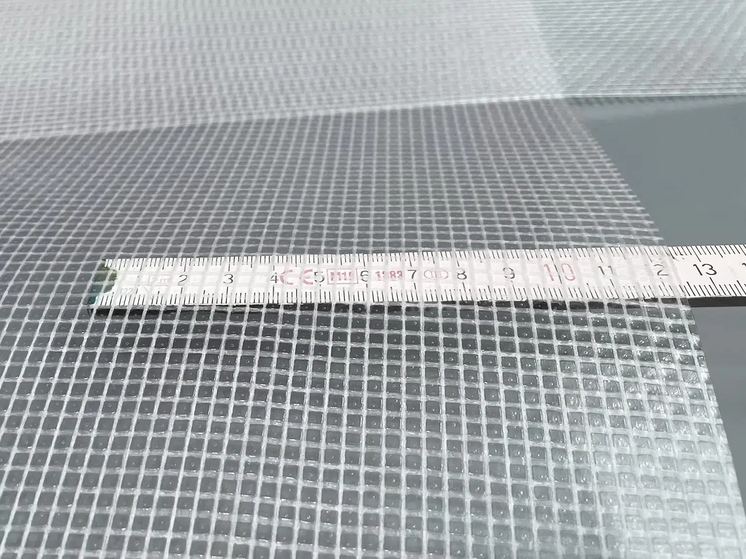 Nahaufnahme eines Maßbandes auf einem gitterförmigen PVC-Netz (550 g/m²) mit kleinen quadratischen Öffnungen. Das 2,18 m breite, hochwertig beschichtete, transluzente PVC-Plane ist mit Maßangaben in Zentimetern abgebildet.