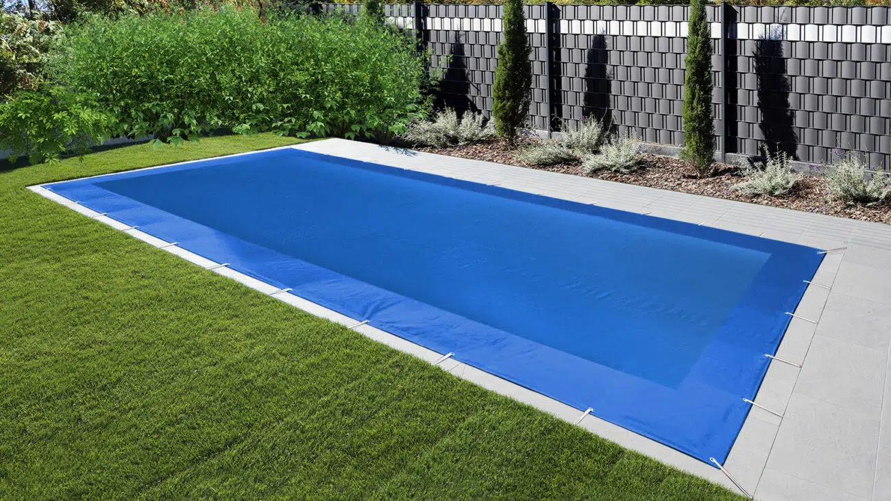 Ein rechteckiger Swimmingpool mit einer blauen rechteckigen Netzabdeckungen liegt zwischen grünem Gras und einem gefliesten Gehweg, während hohe Zäune und dichte Büsche im Hintergrund für Privatsphäre in diesem entspannenden Außenbereich sorgen.