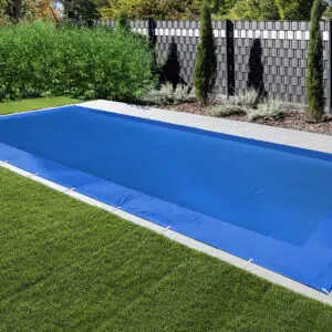 Ein rechteckiger Swimmingpool mit einer blauen rechteckigen Netzabdeckungen liegt zwischen grünem Gras und einem gefliesten Gehweg, während hohe Zäune und dichte Büsche im Hintergrund für Privatsphäre in diesem entspannenden Außenbereich sorgen.