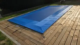 Eine rechteckige Netzabdeckung in Blau ist mit Schnüren fest auf ein Holzdeck im Garten gespannt; im Hintergrund sind Rasen und eine Schubkarre zu sehen.