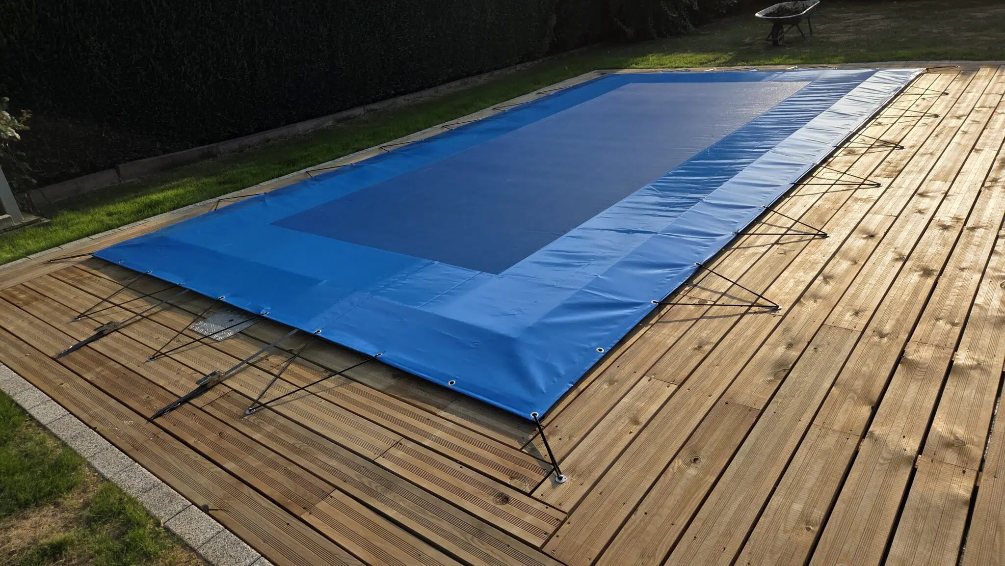 Eine rechteckige Netzabdeckung in Blau ist mit Schnüren fest auf ein Holzdeck im Garten gespannt; im Hintergrund sind Rasen und eine Schubkarre zu sehen.