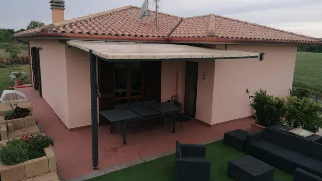 Ein einstöckiges Haus mit Ziegeldach verfügt über eine Terrassendachplane über der Terrasse, die mit Tischen und Stühlen auf Kunstrasen ausgestattet ist und einen Blick auf eine grüne Wiese bietet.