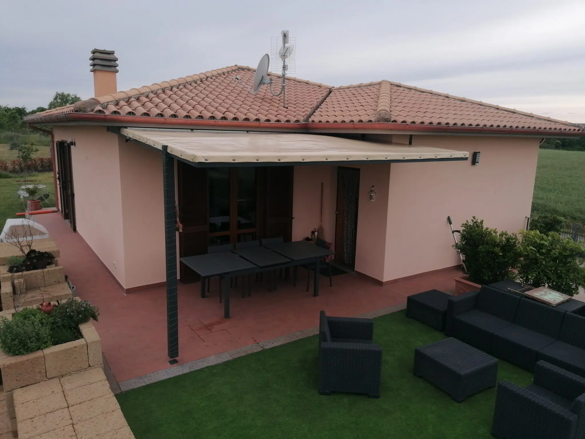 Ein einstöckiges Haus mit Ziegeldach verfügt über eine Terrassendachplane über der Terrasse, die mit Tischen und Stühlen auf Kunstrasen ausgestattet ist und einen Blick auf eine grüne Wiese bietet.