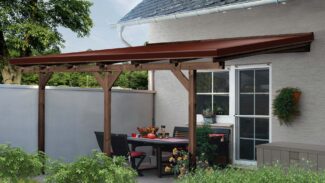 Eine Terrasse mit einer braunen Pergola aus Holz und einer Terrassendachplane an einem Haus mit einem Esstisch, Stühlen, roten Blumen, üppigen grünen Pflanzen und darunter platzierten Sträuchern als Dekoration.