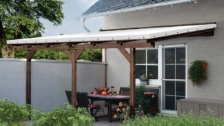 Eine überdachte Außenterrasse mit einer Pergola aus Holz verfügt über eine weiße Terrassendachplane. Darunter ist ein Esstisch mit Stühlen mit roten Kissen mit Blumen und Kürbissen geschmückt, während grüne Sträucher den Vordergrund verschönern.