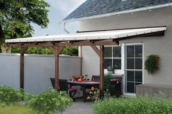 Eine überdachte Außenterrasse mit einer Pergola aus Holz verfügt über eine weiße Terrassendachplane. Darunter ist ein Esstisch mit Stühlen mit roten Kissen mit Blumen und Kürbissen geschmückt, während grüne Sträucher den Vordergrund verschönern.