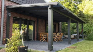 Eine moderne Terrasse präsentiert eine schwarze Pergola mit einer eleganten Terrassendachplane, die mit einem Backsteinhaus verbunden ist. Holzmöbel stehen auf gefliestem Boden, umgeben von üppigem Grün und Topfpflanzen.