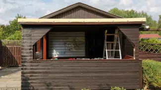 Der Holzschuppen mit großem offenem Fensterrahmen und Leiter im Inneren wird durch eine Terrassendachplane akzentuiert. Er ist von Pflanzen unter einem teilweise bewölkten Himmel umgeben.