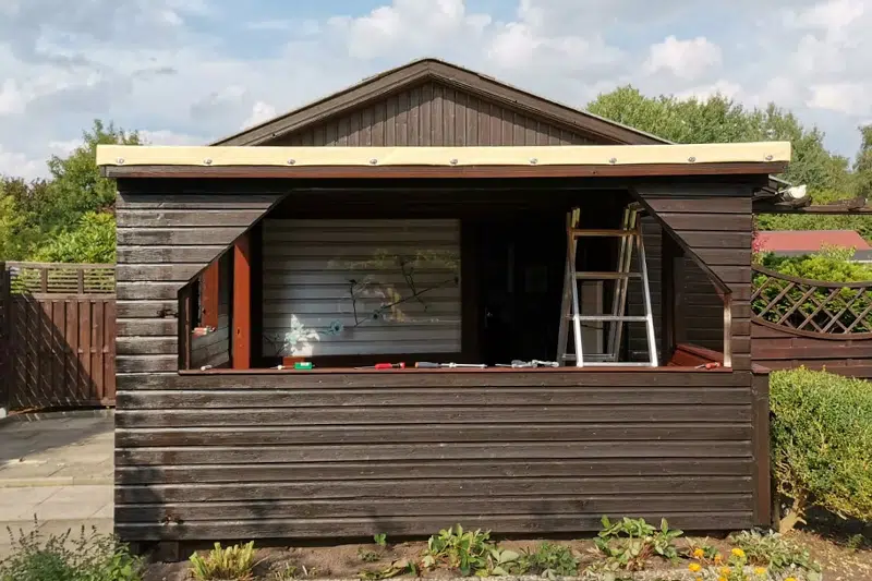 Der Holzschuppen mit großem offenem Fensterrahmen und Leiter im Inneren wird durch eine Terrassendachplane akzentuiert. Er ist von Pflanzen unter einem teilweise bewölkten Himmel umgeben.