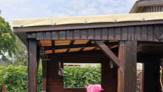 Eine Terrassendachplane schmückt einen hölzernen Pavillon unter bewölktem Himmel. Darunter befinden sich dunkle Holzbalken und Stühle, während im üppigen grünen Vordergrund eine große rosa Blume blüht.