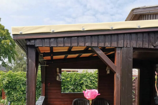 Eine Terrassendachplane schmückt einen hölzernen Pavillon unter bewölktem Himmel. Darunter befinden sich dunkle Holzbalken und Stühle, während im üppigen grünen Vordergrund eine große rosa Blume blüht.