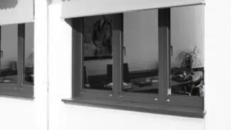 Außenansicht eines Gebäudes mit großen Fenstern und ausgefahrenen Markisen. Im Inneren stehen sichtbare Schreibtische und Büromöbel. Die Markisen sind teilweise ausgerollt und spenden Schatten für die Fenster.