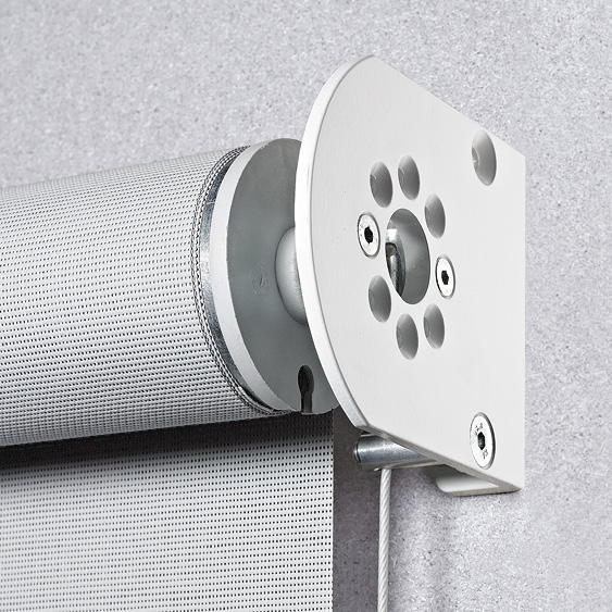 Nahaufnahme eines weißen Rollomechanismus, der an einer grauen Wand montiert ist und dem eleganten Design von Senkrechtmarkisen ähnelt. Die zylindrische Rolle mit perforiertem Design wird durch eine Metallhalterung und eine Zugschnur gesichert.