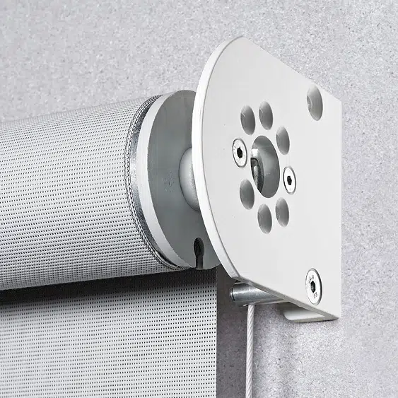 Nahaufnahme eines weißen Rollomechanismus, der an einer grauen Wand montiert ist und dem eleganten Design von Senkrechtmarkisen ähnelt. Die zylindrische Rolle mit perforiertem Design wird durch eine Metallhalterung und eine Zugschnur gesichert.