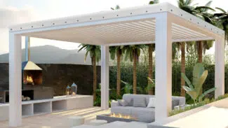 Ein moderner Sitzbereich im Freien verfügt über eine weiße Pergola mit Lamellendach, ein gemütliches Sofa, eine Feuerstelle und einen Grill. Üppige Palmen und eine Bergkulisse runden die ruhige Umgebung ab.