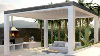 Außenterrasse mit einer modernen Pergola mit verstellbaren Lamellen. Darunter befindet sich ein gemütlicher Sitzbereich mit einer Feuerstelle. Palmen und üppiges Grün umgeben den Raum und im Hintergrund ist ein Außenkamin zu sehen.