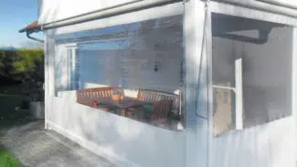 Eine sonnenbeschienene Terrasse hat eine weiße Struktur mit transparenten Vinyl-Einfassungen und eine Senkrechtmarkise-PVC-Plane mit 1x Fenster. Im Inneren stehen auf grauen Pflastersteinen ein Holztisch, Stühle und eine Topfpflanze.