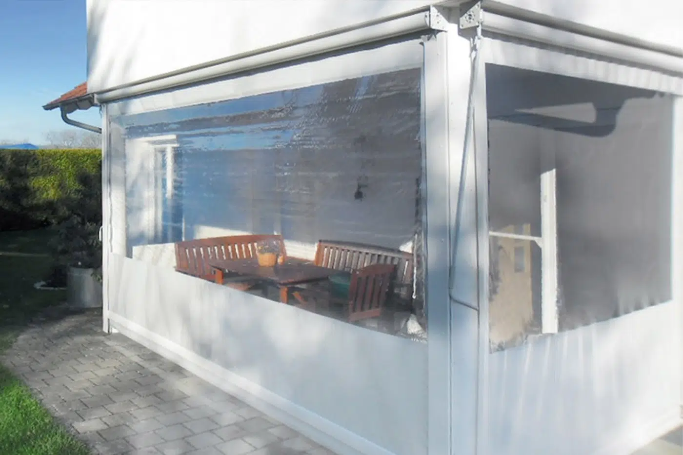 Eine sonnenbeschienene Terrasse hat eine weiße Struktur mit transparenten Vinyl-Einfassungen und eine Senkrechtmarkise-PVC-Plane mit 1x Fenster. Im Inneren stehen auf grauen Pflastersteinen ein Holztisch, Stühle und eine Topfpflanze.