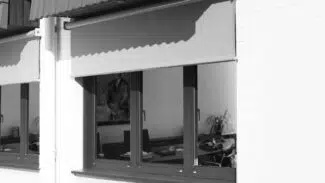 Ein weißes Backsteingebäude hat ein großes, dunkel gerahmtes Fenster mit einer teilweise abgesenkten Senkrechtmarkise PVC-Plane mit 1x Fenster. Im Inneren zeigt der dunkle Raum einen Schreibtisch mit verstreuten Gegenständen und ein Gemälde an der Wand.