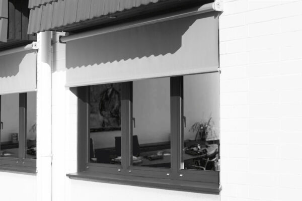 Ein weißes Backsteingebäude hat ein großes, dunkel gerahmtes Fenster mit einer teilweise abgesenkten Senkrechtmarkise PVC-Plane mit 1x Fenster. Im Inneren zeigt der dunkle Raum einen Schreibtisch mit verstreuten Gegenständen und ein Gemälde an der Wand.