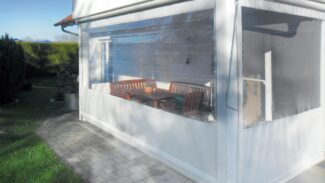 Eine sonnenbeschienene Außenterrasse mit einer senkrecht markierten PVC-Plane mit 1x Fenster umschließt einen Sitzbereich mit einem Holztisch und Bänken neben einem weißen Gebäude, umgeben von Grün und einem gepflasterten Gehweg.