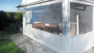 Eine sonnenbeschienene Außenterrasse mit einer senkrecht markierten PVC-Plane mit 1x Fenster umschließt einen Sitzbereich mit einem Holztisch und Bänken neben einem weißen Gebäude, umgeben von Grün und einem gepflasterten Gehweg.