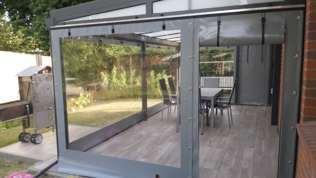 87542219-9548-4c56-ae5e-a5a06dd5a2f2 Eine moderne Außenterrasse verfügt über einen Terrassenplanen-Rollschirm und einen Esstisch. Im Garten steht ein Grill, umgeben von Topfblumen, mit Bäumen und üppigem Grün im Hintergrund.