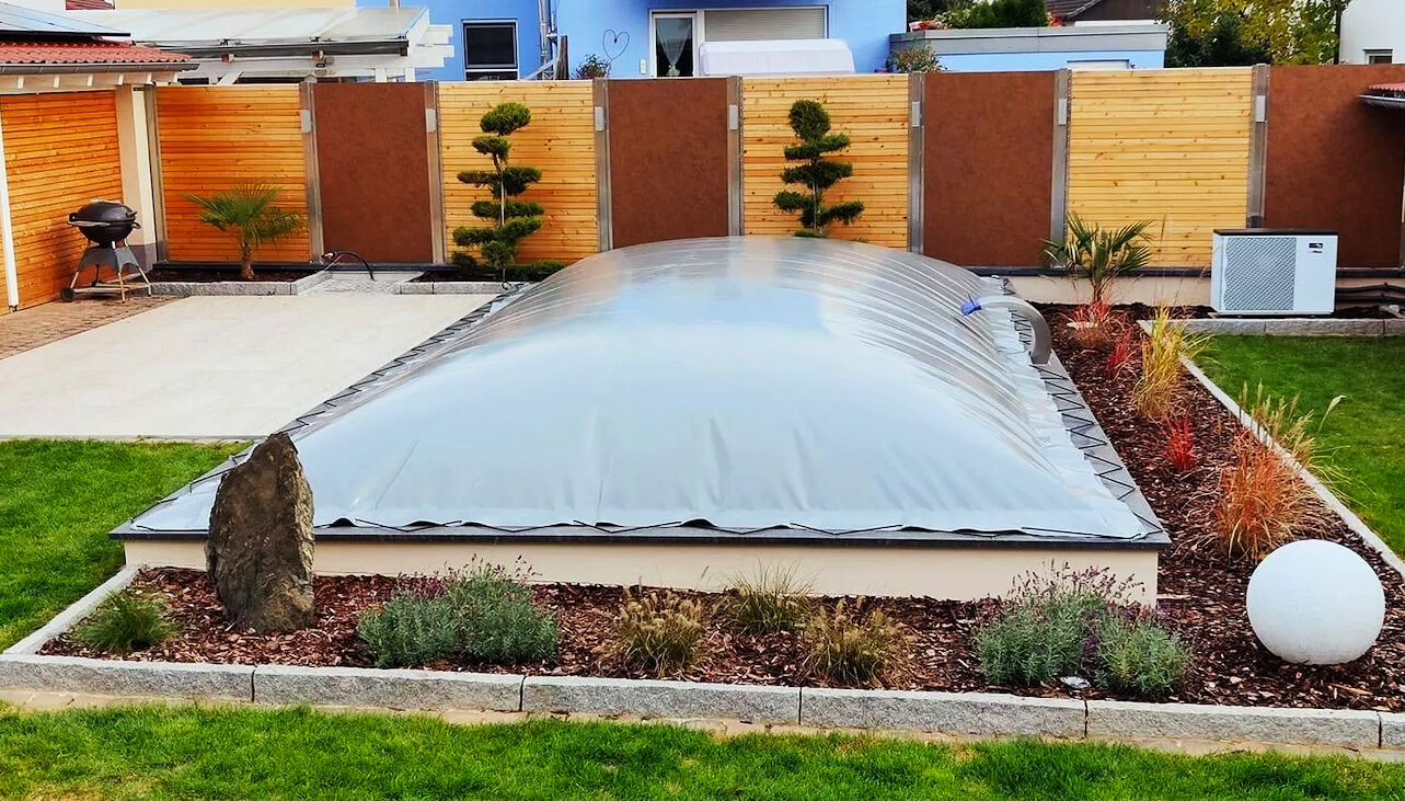 Der Pool im Hinterhof, der von einer aufblasbaren Poolplane-Kuppel umschlossen ist, ist von einer gepflasterten Terrasse, üppigem Gras und Landschaftspflanzen umgeben. Eine runde weiße Skulptur ziert die rechte Seite; links neben dem blauen Haus steht ein Grill.