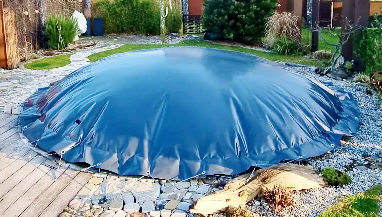 In einer Hinterhofszene ist ein Poolplane zu sehen, eine schwarze, kuppelförmige Poolabdeckung, umgeben von Felsen und einem Holzdeck. Der Bereich ist mit verschiedenen Pflanzen und Sträuchern bepflanzt, im Hintergrund sind Häuser zu sehen.