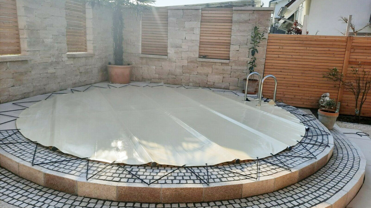 Ein runder Pool, der mit einer Poolplane abgedeckt ist, ruht auf einer runden, erhöhten Steinplattform mit Stufen. Eine Topfpalme und eine Pflanze schmücken den Bereich, der von einem Stein- und Holzzaun im sonnendurchfluteten Innenhof umgeben ist.