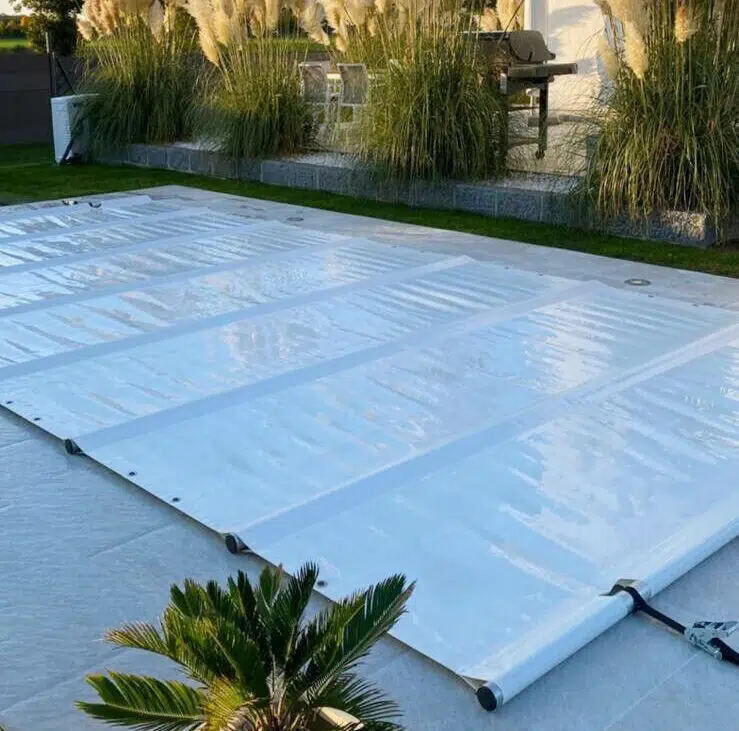 Eine Poolplane deckt den Swimmingpool in einer Gartenumgebung ab, wobei auf der Terrasse vor dem Hintergrund eines klaren Himmels hohe Ziergräser und eine kleine Topfpflanze sichtbar sind.