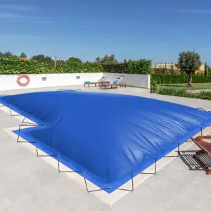 Ein Swimmingpool mit aufblasbaren Poolplanen in L-Form, umgeben von einer gefliesten Terrasse. In der Nähe befinden sich zwei Holzliegen und ein Rettungsring, eingebettet in eine grüne Umgebung und farbenfrohe Gebäude.
