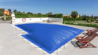 Ein Swimmingpool mit aufblasbaren Poolplanen in L-Form, umgeben von einer gefliesten Terrasse. In der Nähe befinden sich zwei Holzliegen und ein Rettungsring, eingebettet in eine grüne Umgebung und farbenfrohe Gebäude.
