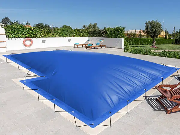 Eine blaue Poolplane-Abdeckung ist sicher über einem Pool auf einer sonnigen Terrasse gespannt. Liegestühle, Grünpflanzen und eine weiße Wand mit einem Rettungsring umgeben die Szene unter dem klaren blauen Himmel.