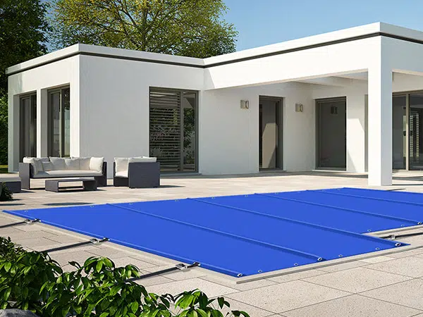 Das moderne Haus verfügt über eine geräumige Terrasse mit einer straff gespannten blauen Poolplane, die den Pool bedeckt. Weiße Wände und große Glasfenster ergänzen das weiße Sofa und den kleinen dunklen Tisch, während üppiges Grün die Szenerie akzentuiert.
