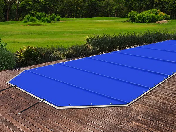 Eine leuchtend blaue Poolplane bedeckt einen rechteckigen Pool auf einem Holzdeck. Im Hintergrund erstreckt sich unter dem klaren Himmel eine üppig grüne Landschaft mit Bäumen und Sträuchern.