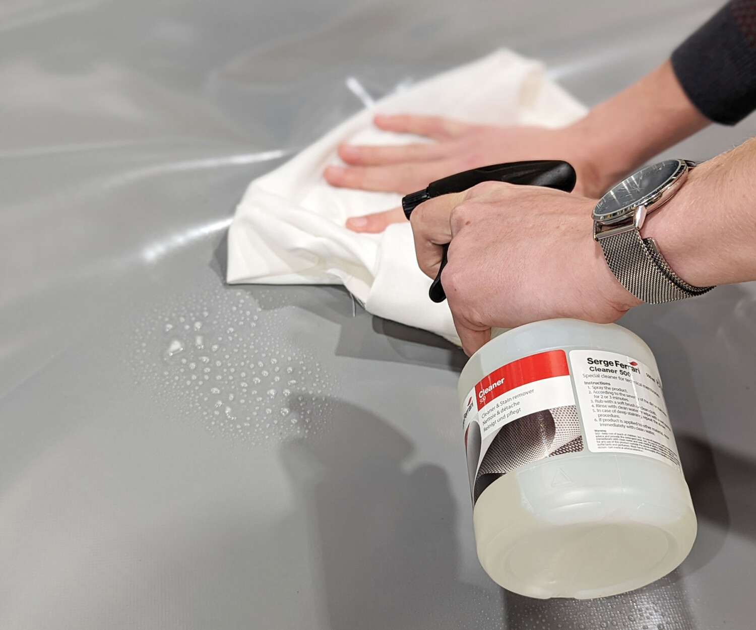 Person reinigt eine graue Oberfläche mit einem weißen Tuch und einer Sprühflasche. Auf der Oberfläche sind Blasen sichtbar. Die Person trägt eine Uhr und das Bild konzentriert sich auf die Reinigungsaktion.