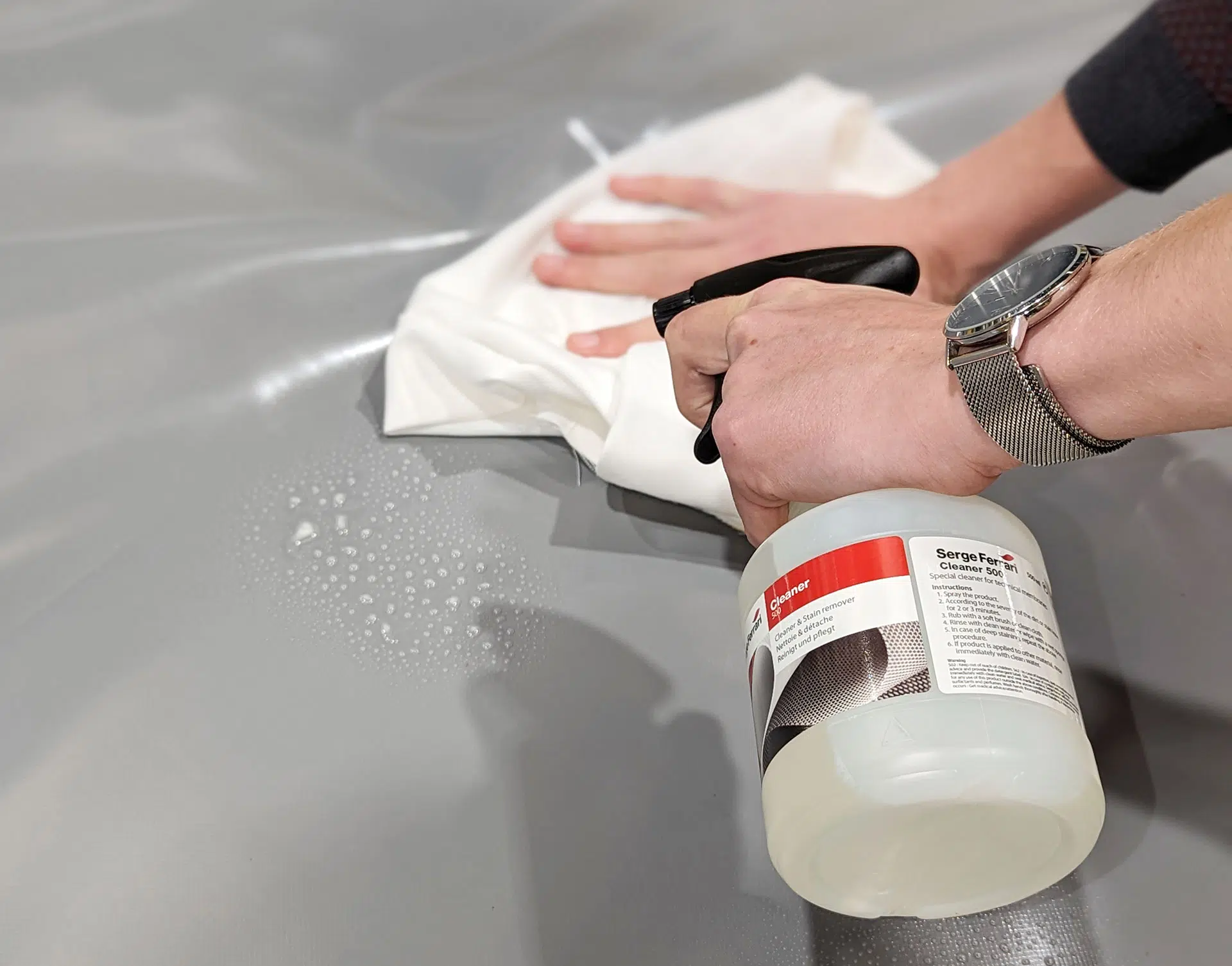 Person reinigt eine graue Oberfläche mit einem weißen Tuch und einer Sprühflasche. Auf der Oberfläche sind Blasen sichtbar. Die Person trägt eine Uhr und das Bild konzentriert sich auf die Reinigungsaktion.