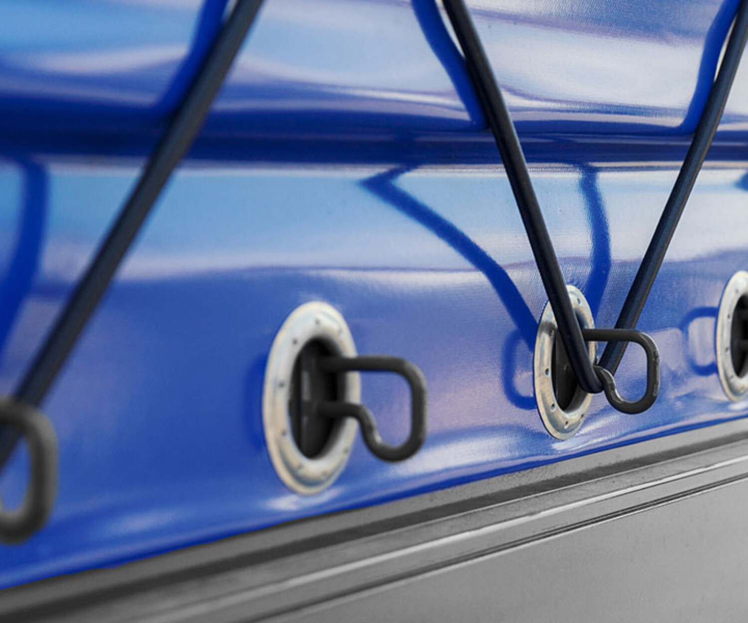 Nahaufnahme einer blauen Plane, die mit elastischen Schnüren und Haken befestigt ist. Der Stoff ist straff gespannt, und die Reflexionen lassen auf eine glatte Oberfläche schließen. Die Metallringe sind gleichmäßig entlang der Kante angeordnet und betonen die Präzision.