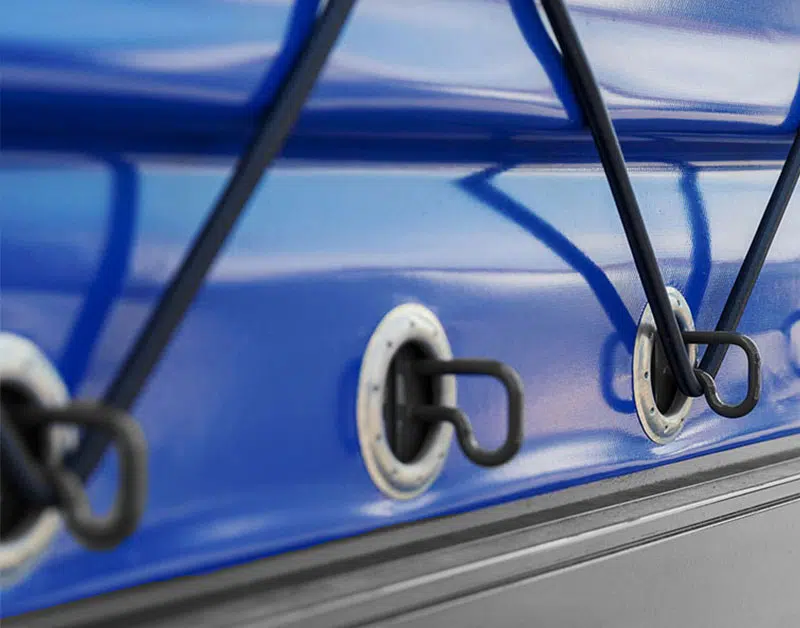 Nahaufnahme einer blauen LKW-Seite mit elastischen Kordeln, die durch Metallösen gehakt sind. Die diagonale Anordnung der schwarzen Kordeln kontrastiert mit der blauen Oberfläche und erzeugt ein Muster auf der glatten Oberfläche.