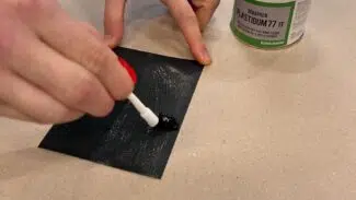 Eine Hand trägt mit einem Pinsel Klebstoff aus einem 750-ml-Gebinde PVC-Kleber - Plastigum77 - auf eine schwarze rechteckige Fläche auf.