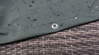 Nahaufnahme einer dunkelgrünen Plane mit Wassertropfen auf der Oberfläche, die über einer gewebten Rattanoberfläche liegt. Nahe dem Rand der Plane ist eine Metalltülle sichtbar.