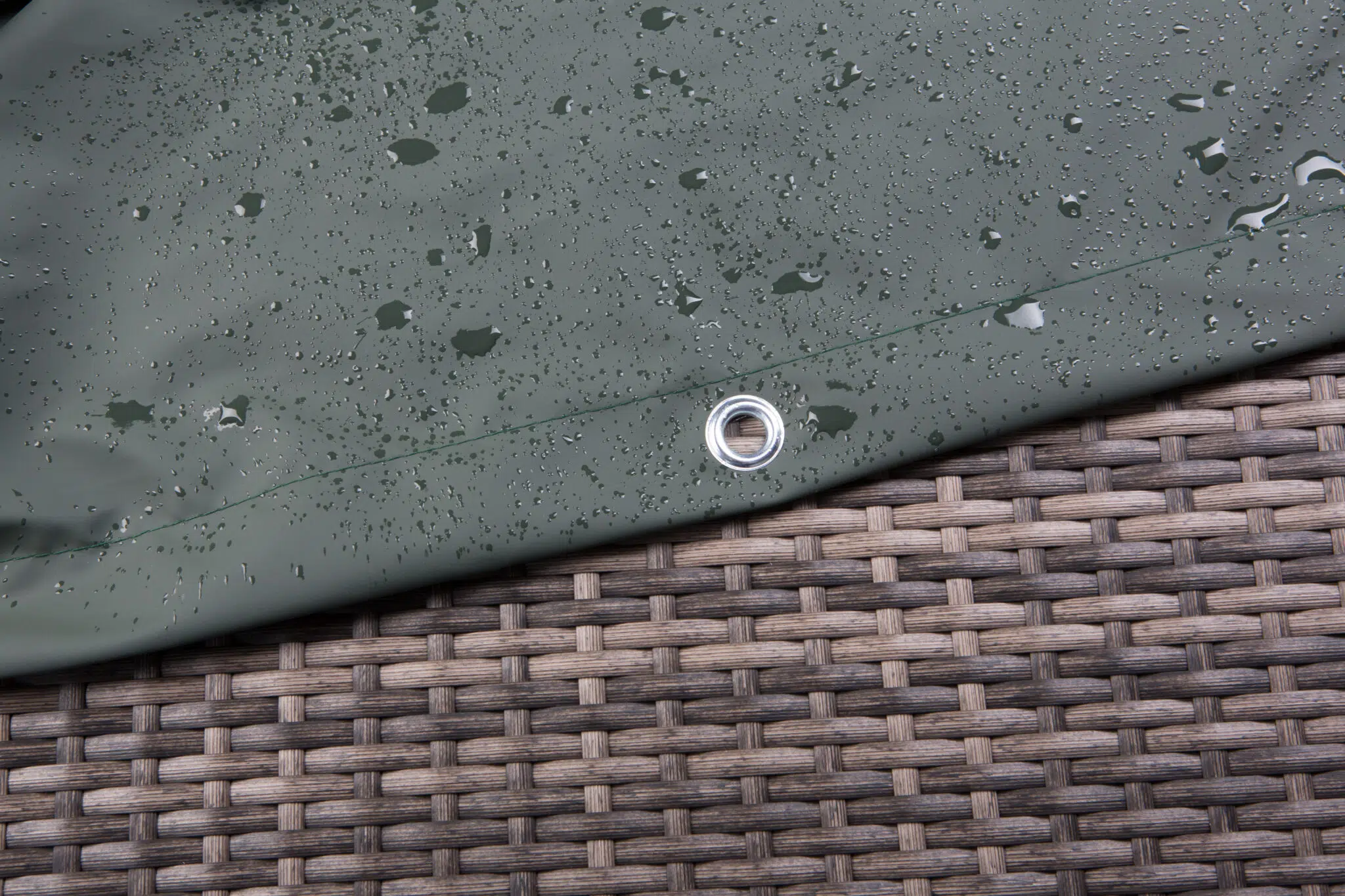 Nahaufnahme einer dunkelgrünen Plane mit Wassertropfen auf der Oberfläche, die über einer gewebten Rattanoberfläche liegt. Nahe dem Rand der Plane ist eine Metalltülle sichtbar.