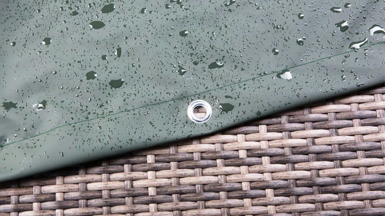 Nahaufnahme einer grünen Plane, die oft für Abdeckhauben verwendet wird, mit Wassertropfen und einer Metallöse, die auf einer gewebten Rattanoberfläche ruht. Die Tropfen sind über die Plane verstreut, was darauf hinweist, dass sie vor Kurzem nass geworden ist.