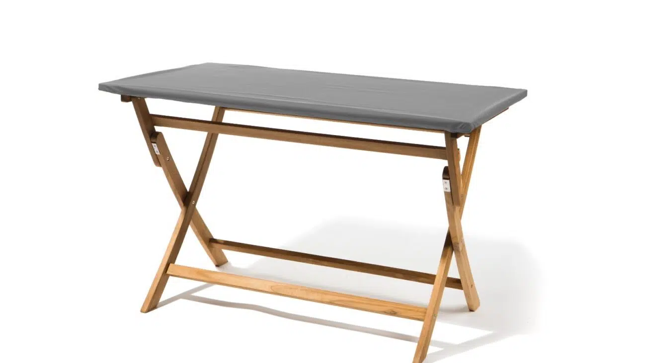 Ein Holzklapptisch mit grauer Tischplatte und X-Rahmendesign wirkt auf einem weißen Hintergrund tragbar, aufgewertet durch eine eckige Tischplattenhaube mit Gummizug (Teak Safe) für zusätzlichen Stil.