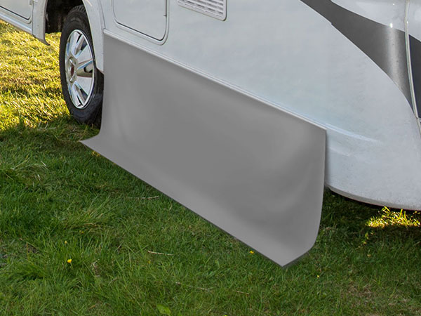 Eine graue Wohnmobil-Windschürze, unverzichtbar fürs Camping, ist an der Seite eines auf Gras geparkten Wohnmobils angebracht. Sie liegt bis zum Boden auf und bietet sowohl Isolierung als auch Schutz vor den Elementen.