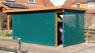 Ein Carport mit Seitenteilen aus blaugrünem Stoff, einschließlich einer Carport- oder Terrassenplane aus Netzgewebe, bietet einem geparkten Auto in einem Wohngebiet mit modernen Häusern und eingezäunten Gärten teilweise Schutz.
