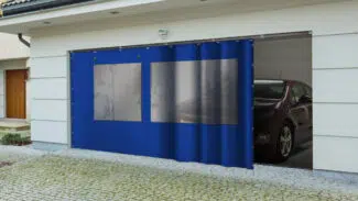 Eine moderne Garage zeigt eine blaue Schiebeplane mit 2x Fenster/ mit 1x Tür (mit Fenster), die teilweise geöffnet ist und in der ein Auto auf einer Kopfsteinpflasterauffahrt steht.