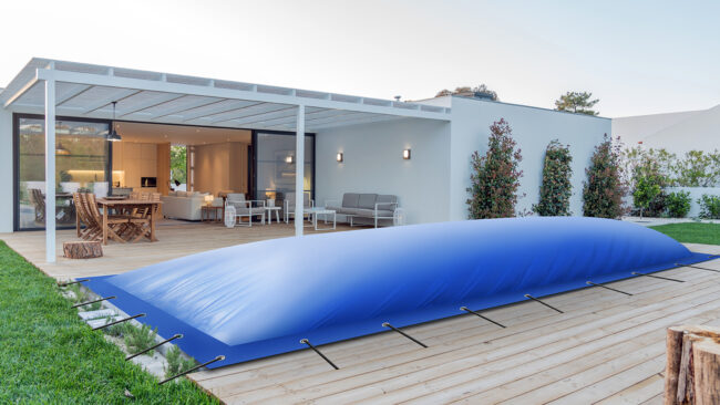 Ein modernes Haus mit einer Holzterrasse und einer blauen Poolplanen-Abdeckung, die über dem rechteckigen Pool aufgeblasen ist; Sitz- und Essbereiche im Freien befinden sich unter einer Pergola.