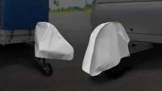 Zwei Anhängerkupplungen, die jeweils mit einer weißen Schutzabdeckung versehen sind, werden auf einer gepflasterten Fläche neben einem Anhänger und einem Wohnmobil abgestellt.